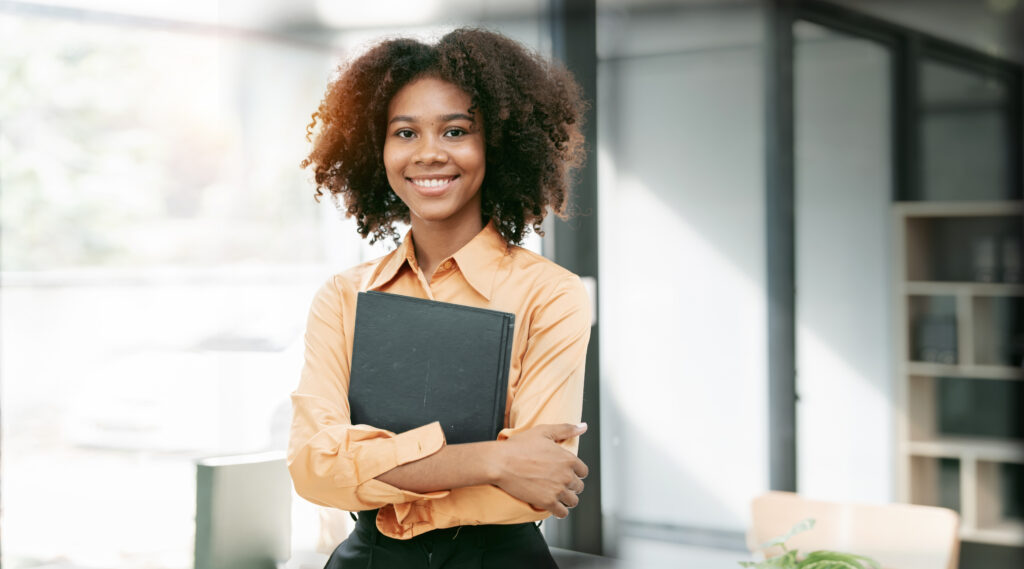 A young professional holding a book and standing cheerfully in a bright office setting.